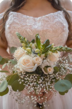 Bridal bouquet with cream roses, baby's breath, and eucalyptus against white lace dress.