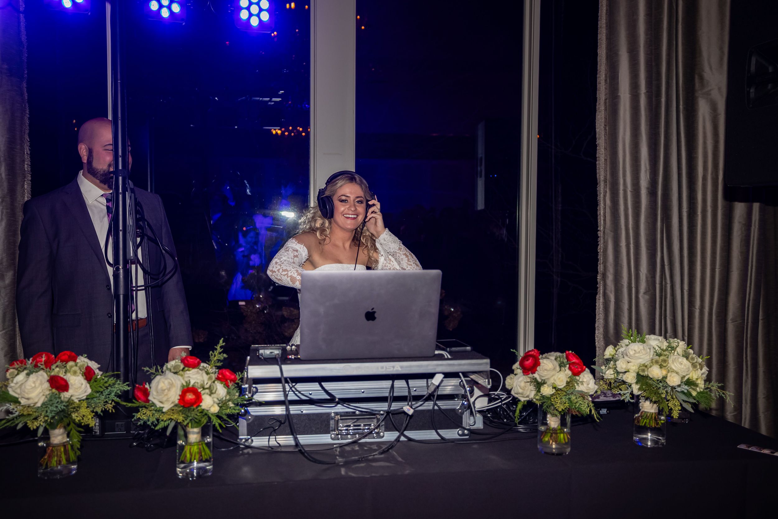 DJ plays music at a wedding reception with blue lighting and floral decorations on tables.
