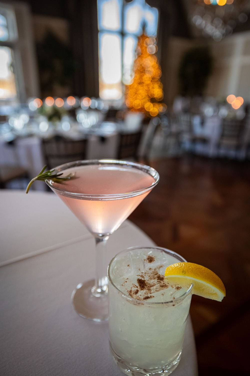 Two elegant cocktails displayed on a white tablecloth with a blurred Christmas tree glowing in the background.