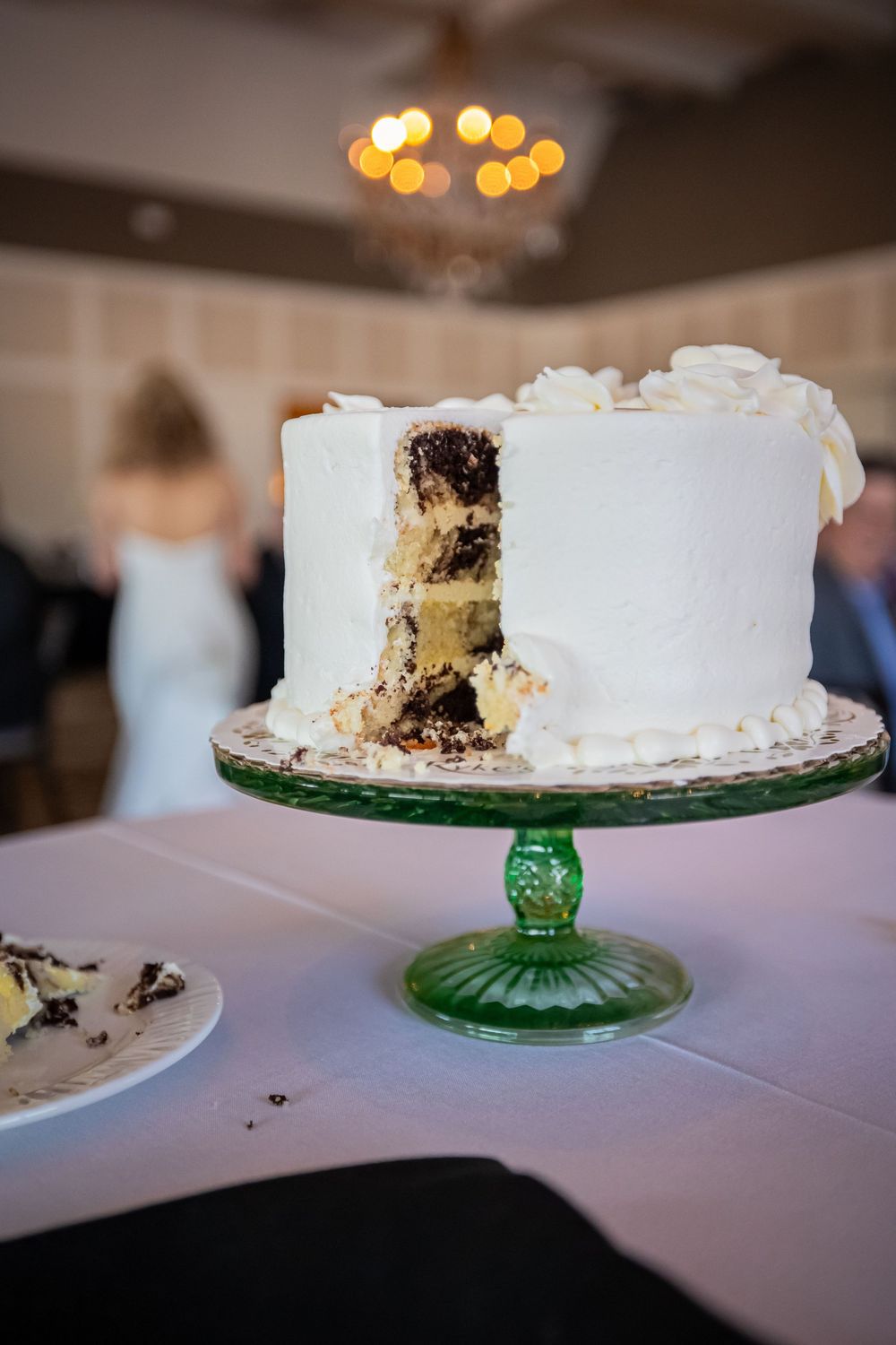 A photo showing a white wedding cake after being cut and served on a green glass cake stand with bokeh lights in background.