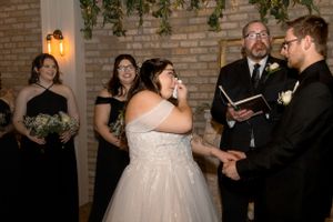 Couple embraces during indoor wedding ceremony in front of exposed brick wall with hanging greenery and lanterns.