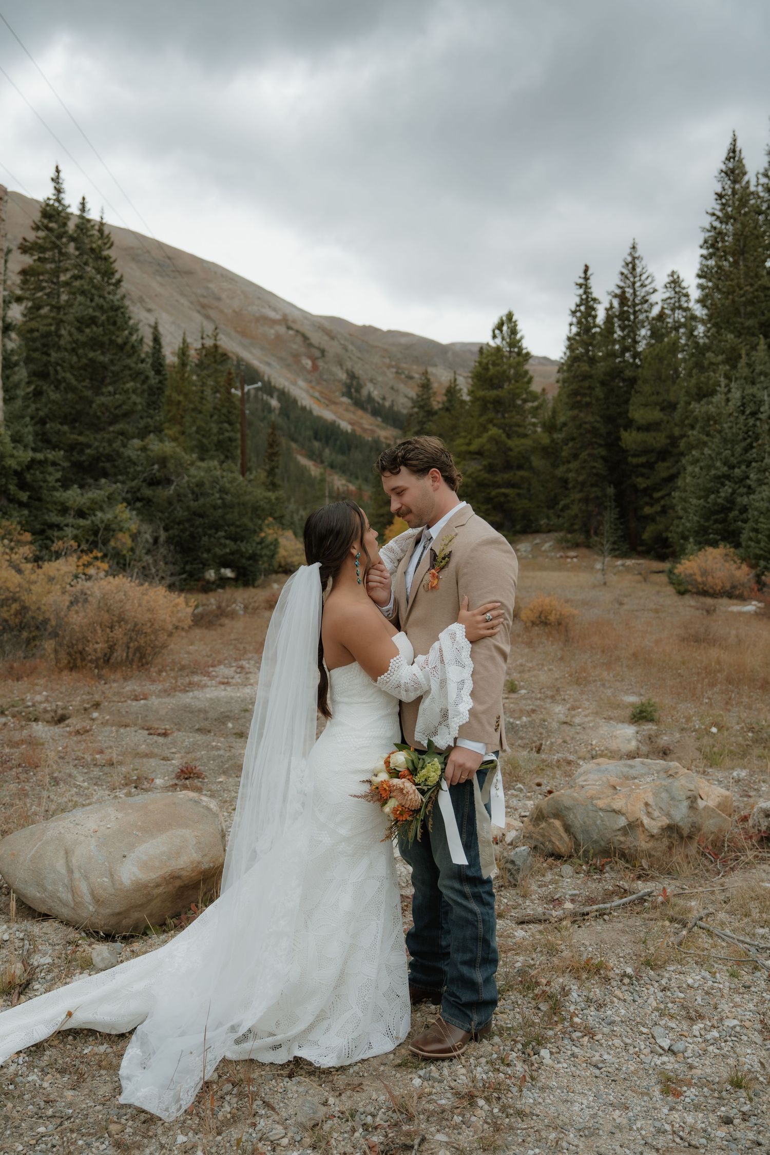 Couple in wedding attire standing by mountain lake with snow-capped Maroon Bells peaks.