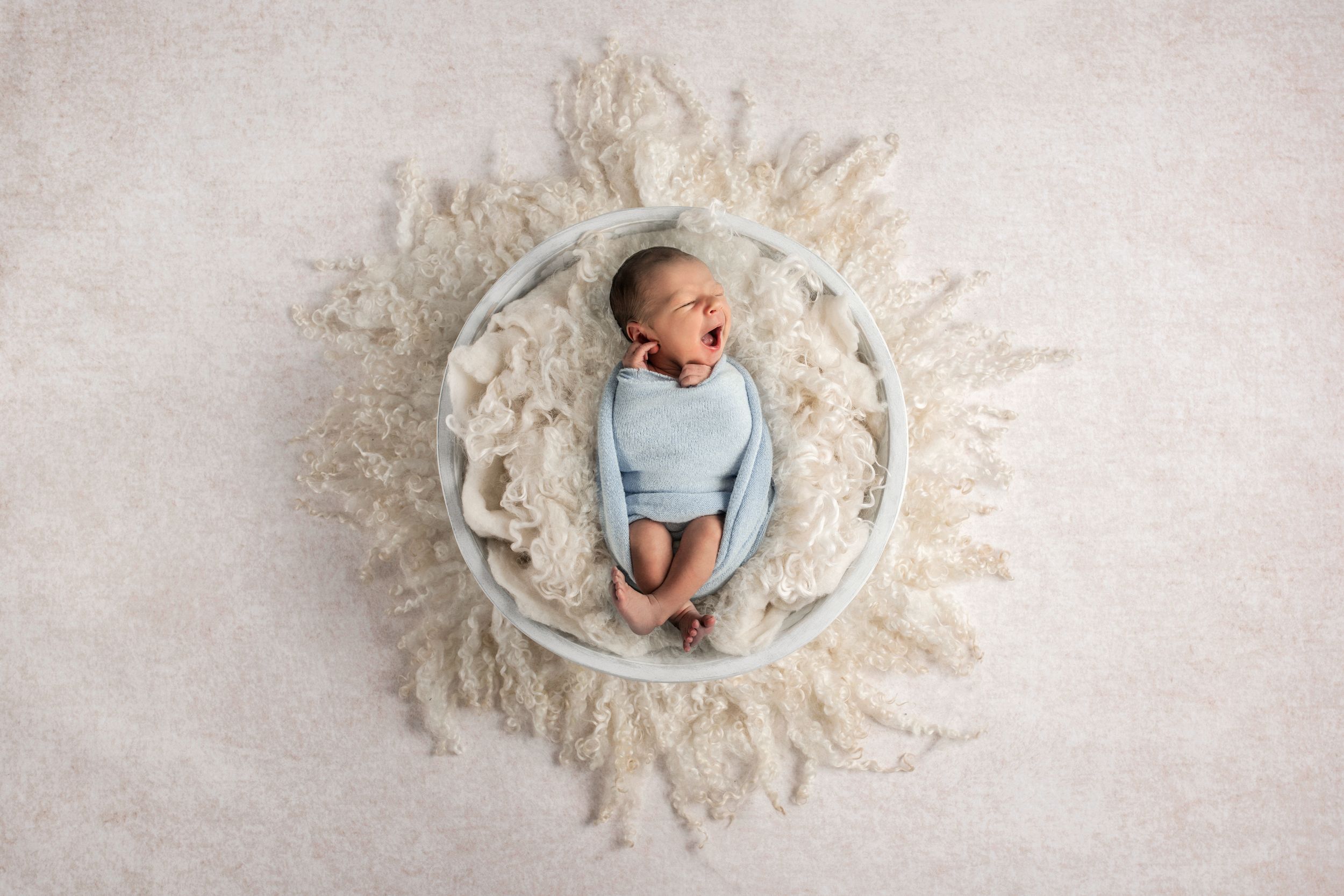 Newborn baby in a light blue outfit yawning while lying in a round bowl with cream fluffy texture for Marietta newborn photographer.