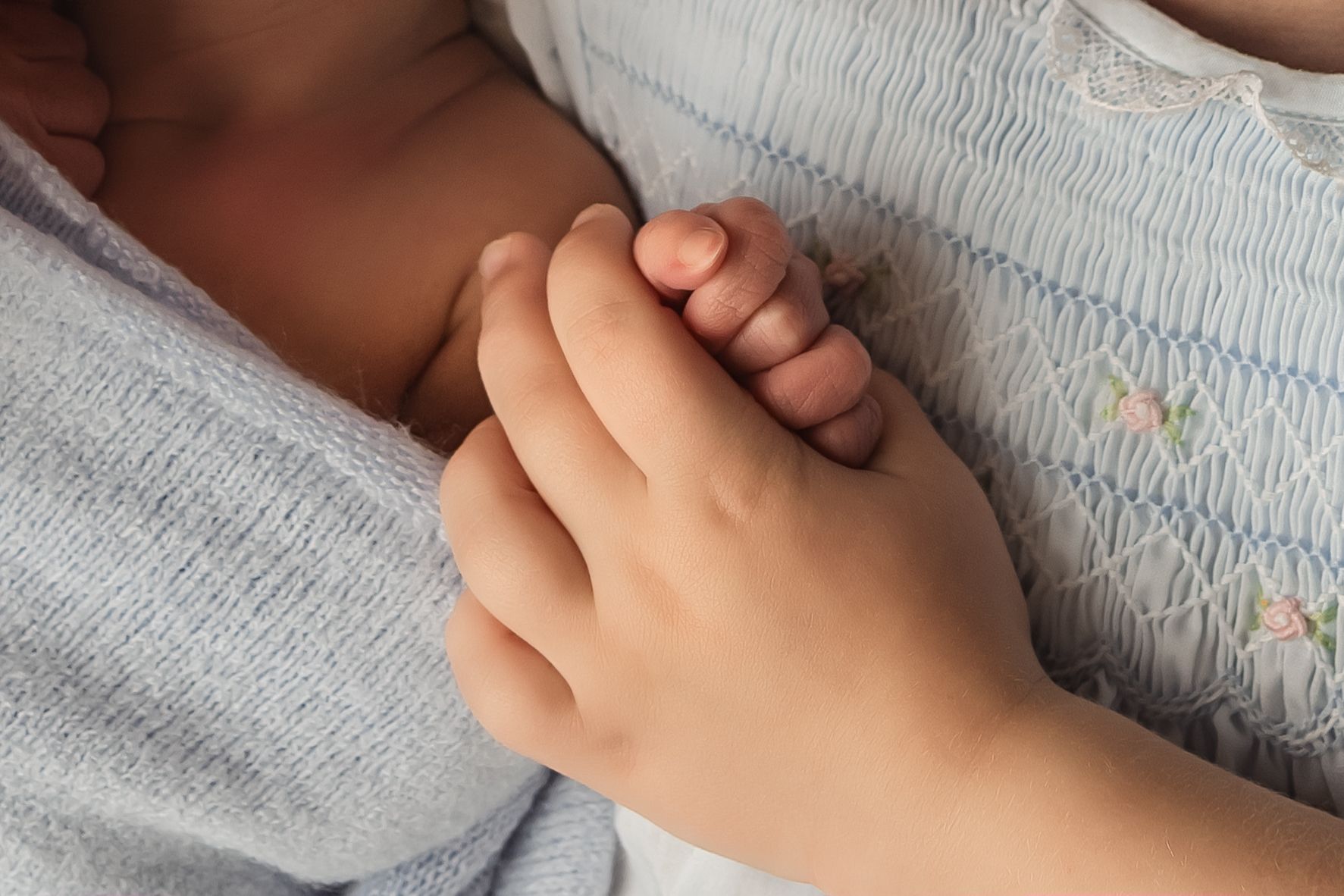 A child's hand gently holding a newborn baby's tiny fingers, wrapped in soft blue blankets.