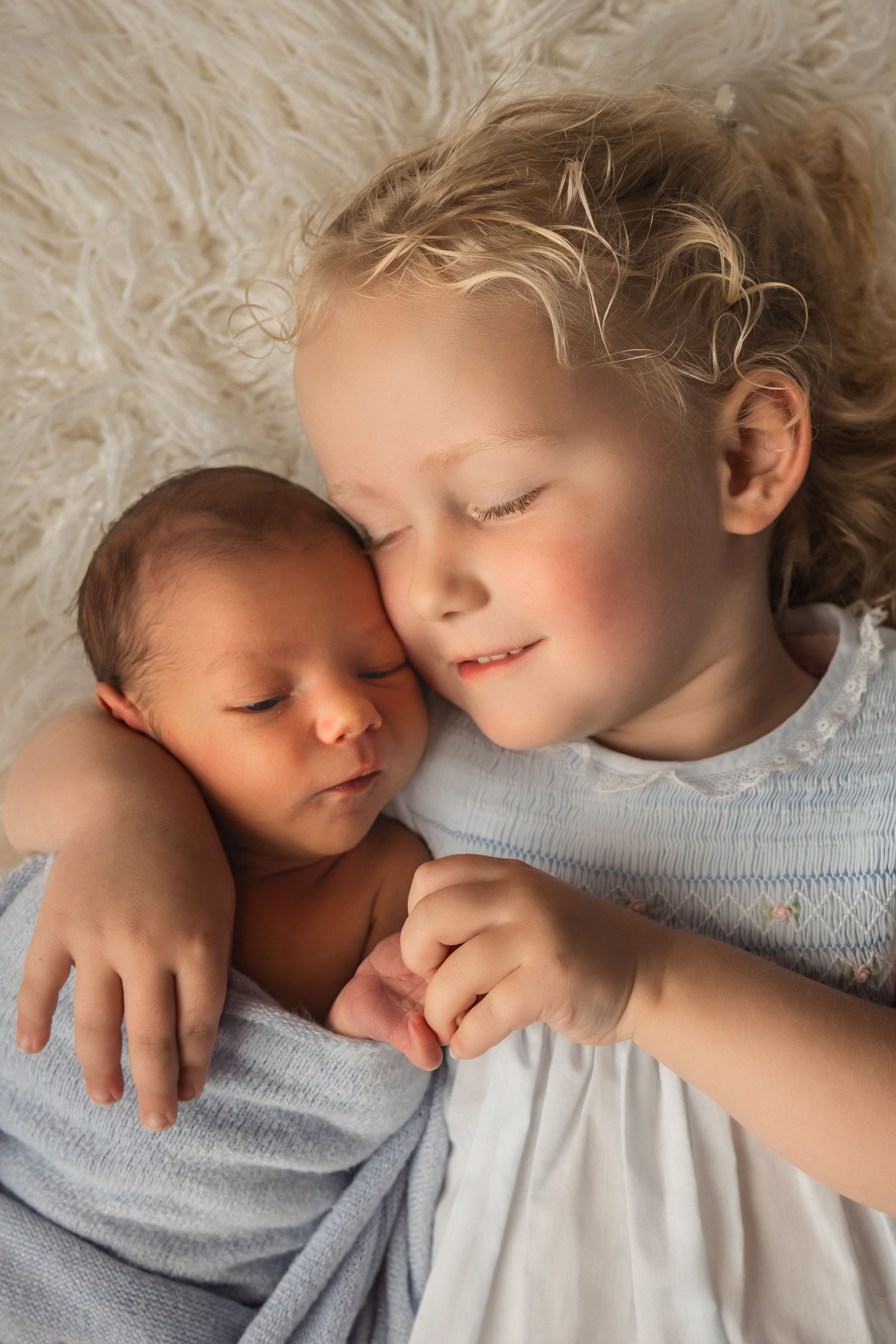 Blonde toddler girl in white dress cuddles sleeping newborn baby sibling wrapped in blue on fluffy white blanket.