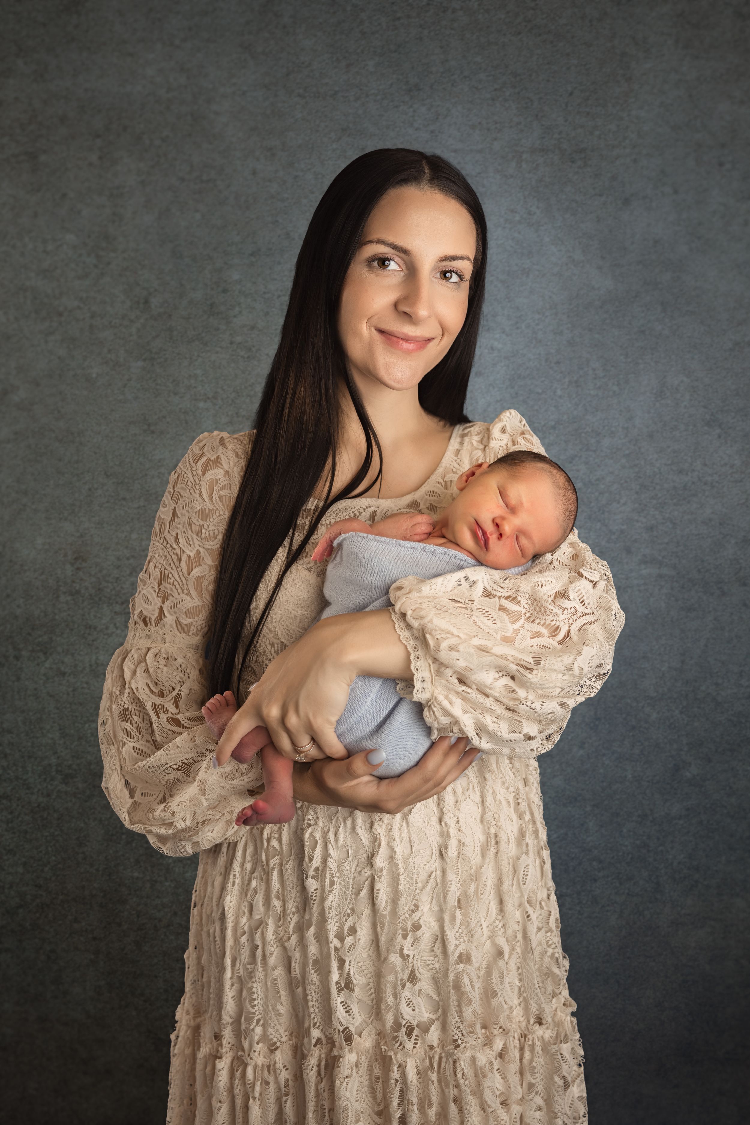 Young mother in lace dress tenderly holds sleeping newborn baby against gray studio background.