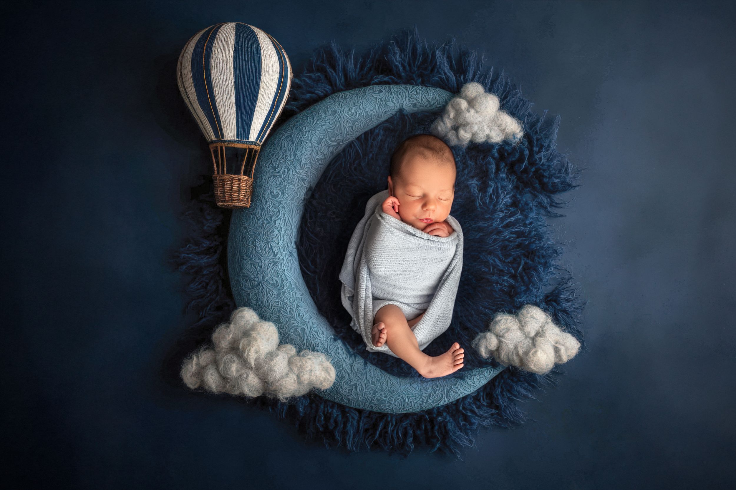 Newborn baby sleeping on a blue crescent moon prop with clouds and a hot air balloon in a studio photo by marietta newborn photographer