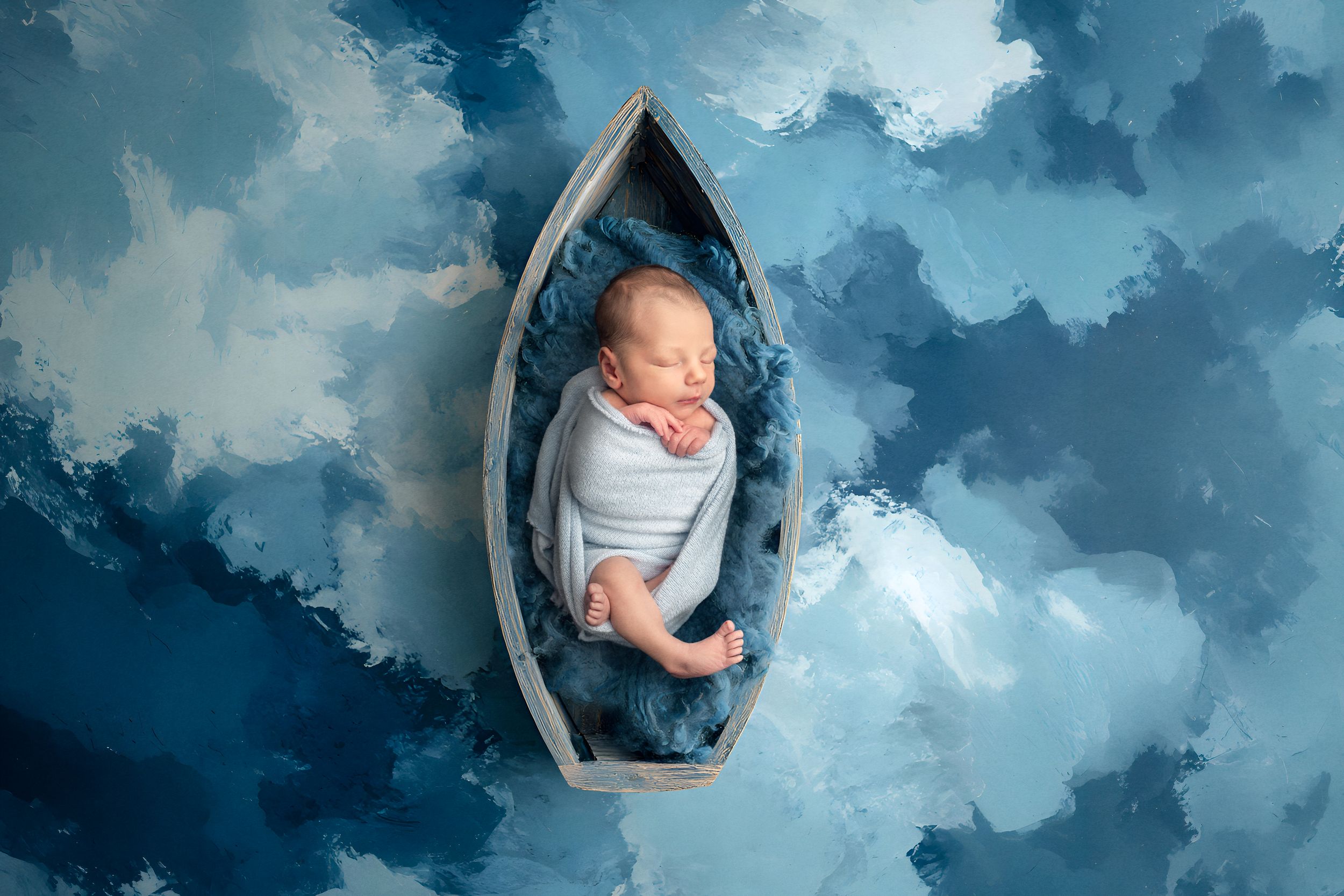 Newborn baby swaddled in blue sleeping peacefully in a small wooden boat on a blue painted background for studio newborn photo session.