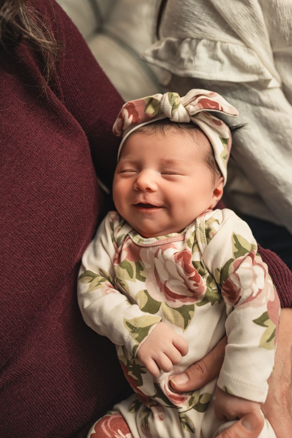 A smiling newborn baby girl wearing a floral outfit and matching headband rests in her mother's arms for newborn photo shoot.