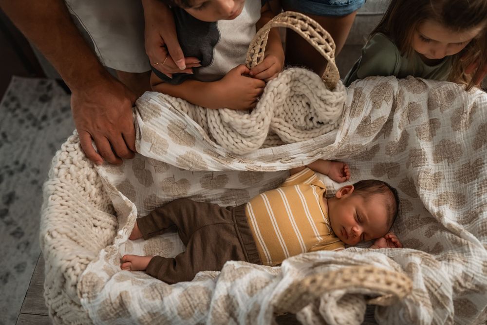 A sleeping newborn baby wearing a striped onesie lies nestled in soft cream-colored lace blankets while family looks down from above.