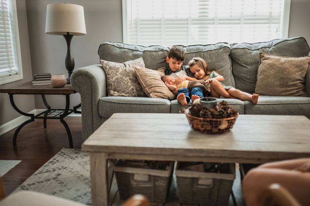 Big brother and sister holding newborn baby brother on a gray couch in a cozy living room with a coffee table and lamp.
