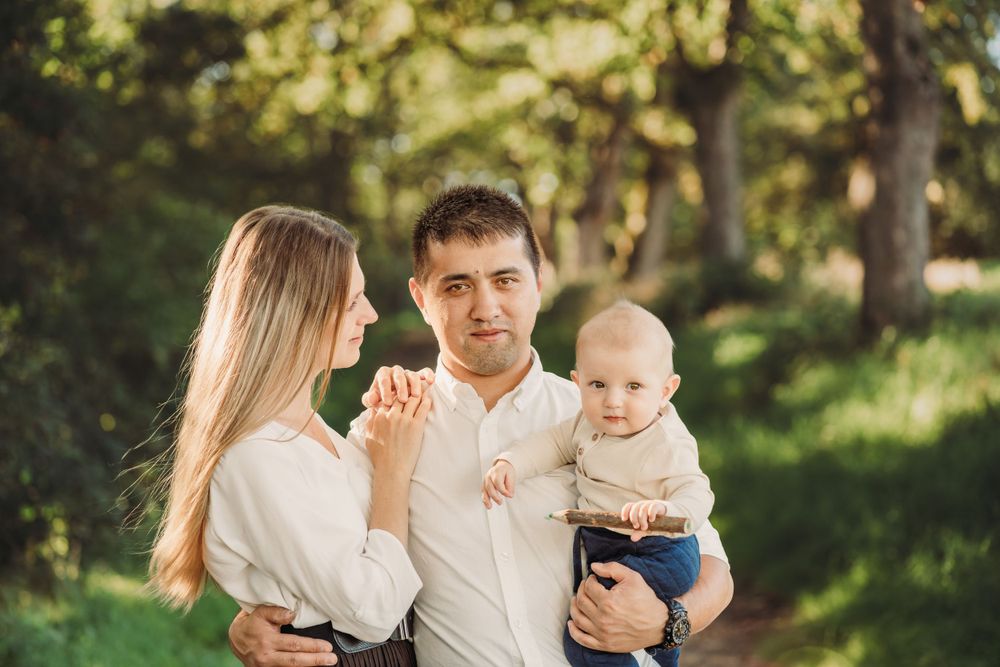 A couple shares a tender moment while holding their baby in a sunlit forest setting.