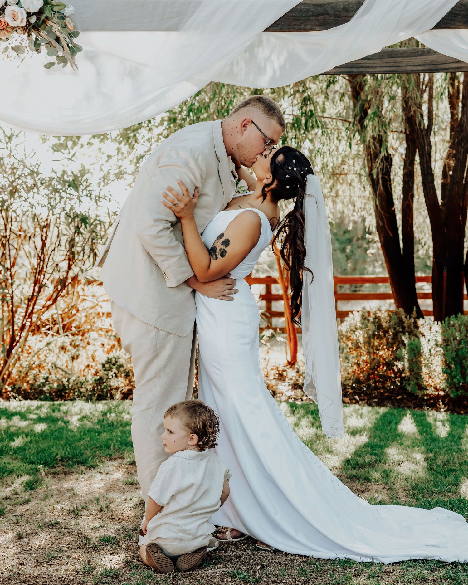 Sweet moment at an outdoor garden wedding with bride in white gown and two others in beige suits under draped white fabric.