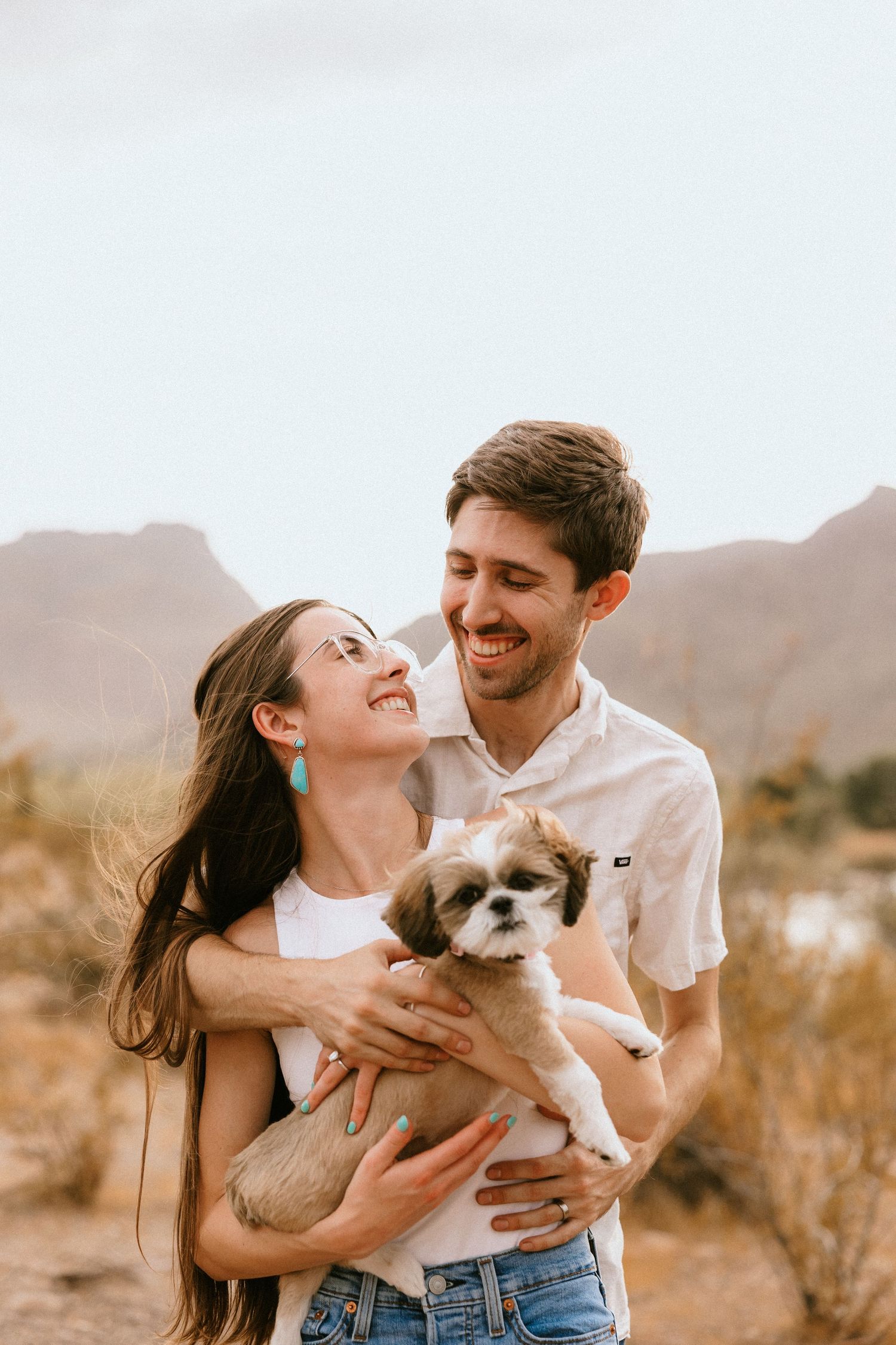 A happy couple holds their small dog while posing in the desert landscape.
