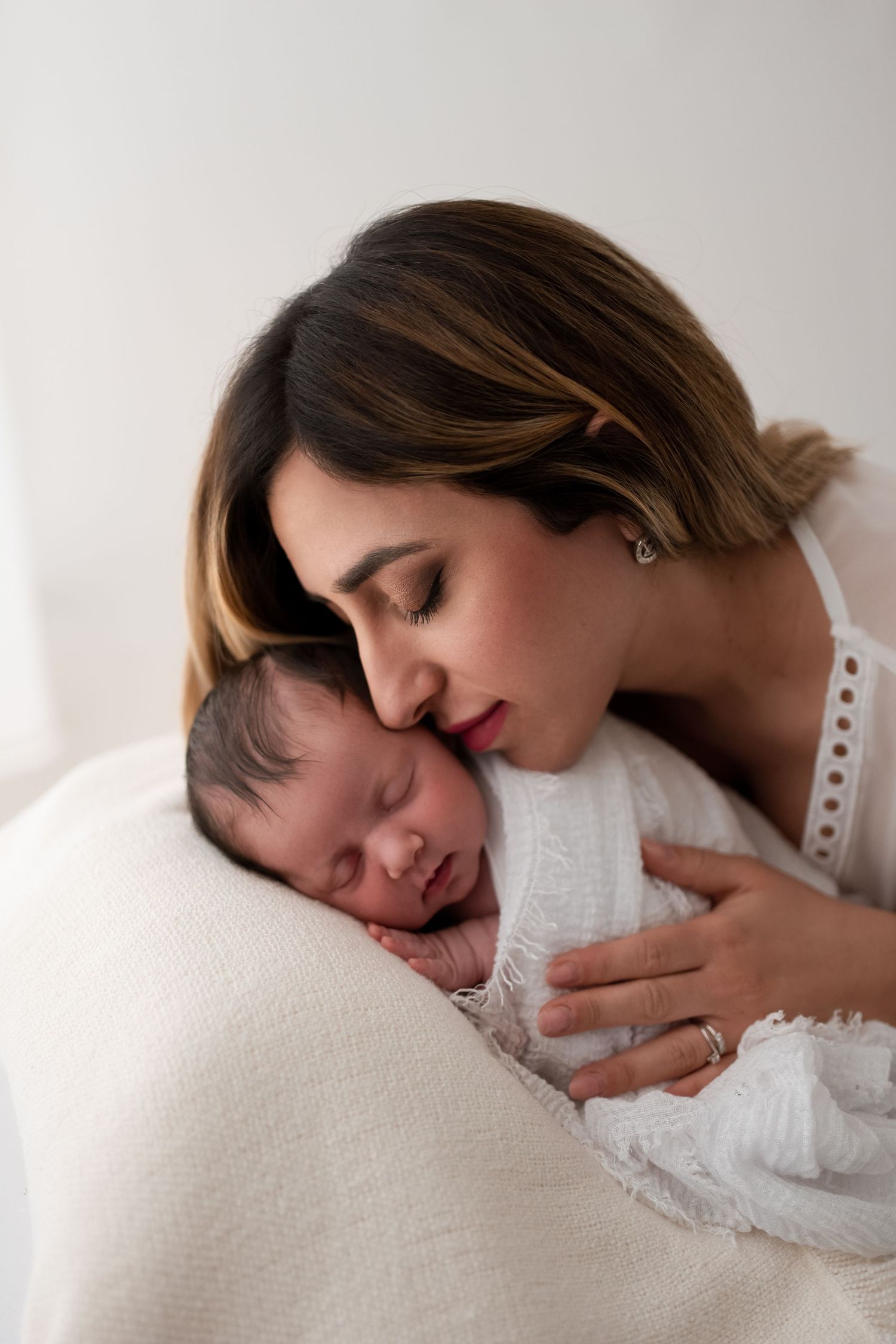 Tender mother leaning over her sleeping newborn baby wrapped in white muslin cloth.