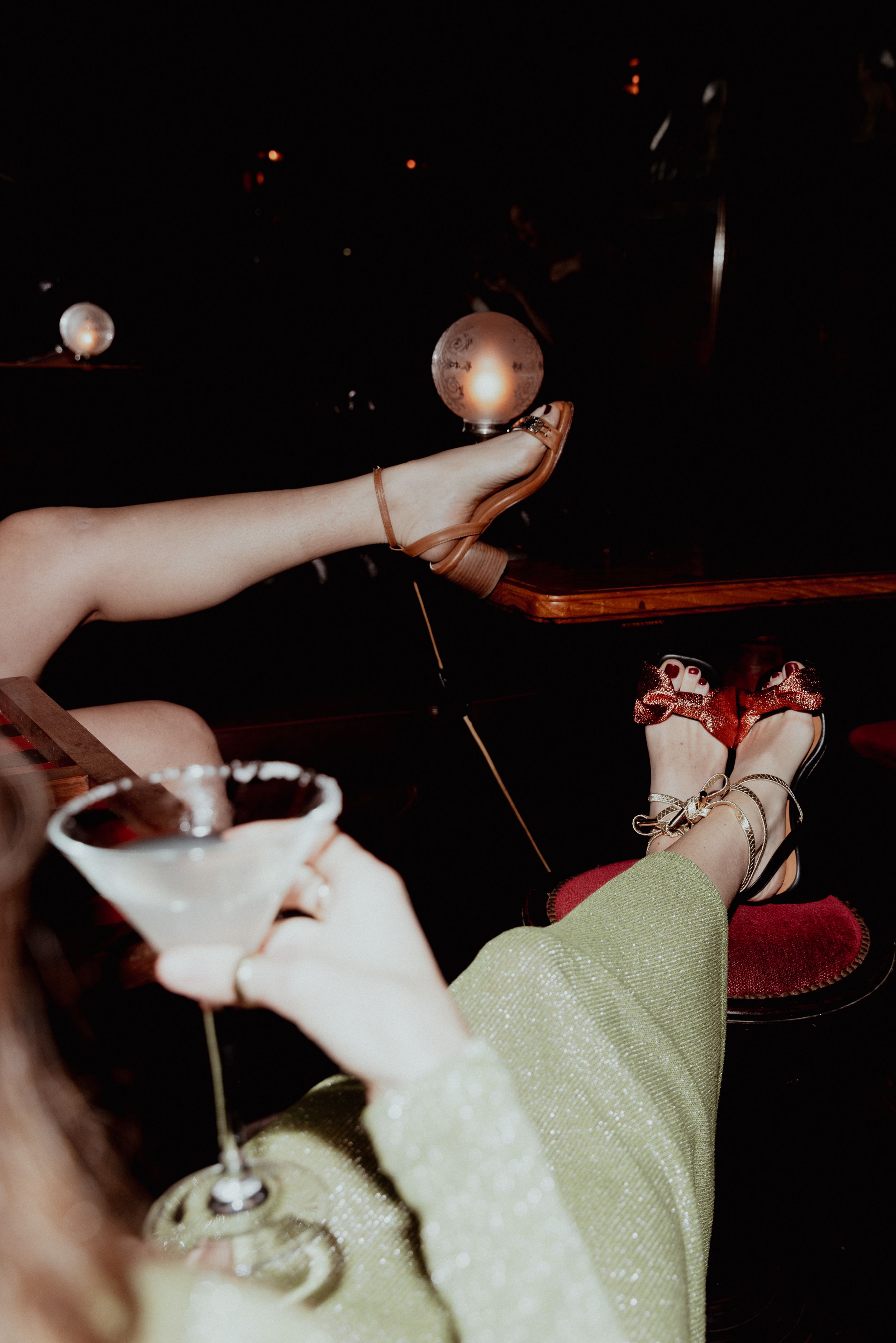 Legs in sparkly dress and red heels propped up with cocktail glasses on a dark table.