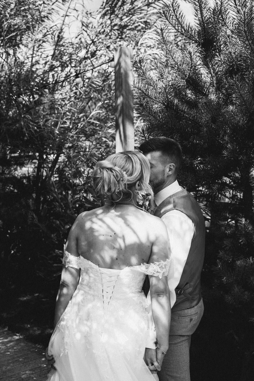 A candid black and white shot of the bride and groom sharing a private moment beneath dappled sunlight in a forest
