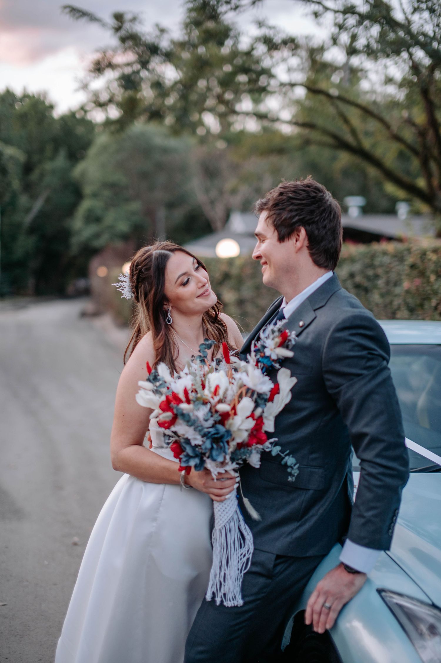 Couple shares romantic moment by car with red and white bouquet at sunset.