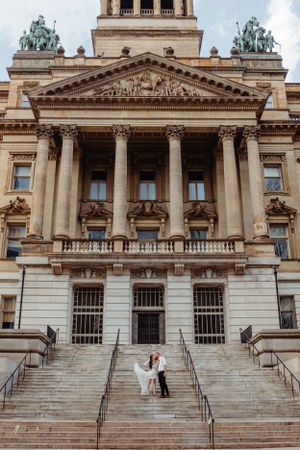 Wedding couple poses on steps of a grand neoclassical building with ornate architectural details.