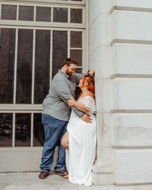 Couple in casual and formal attire embracing against an architectural wall.
