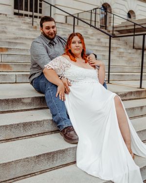 Couple in wedding attire sitting casually on stone steps of building.