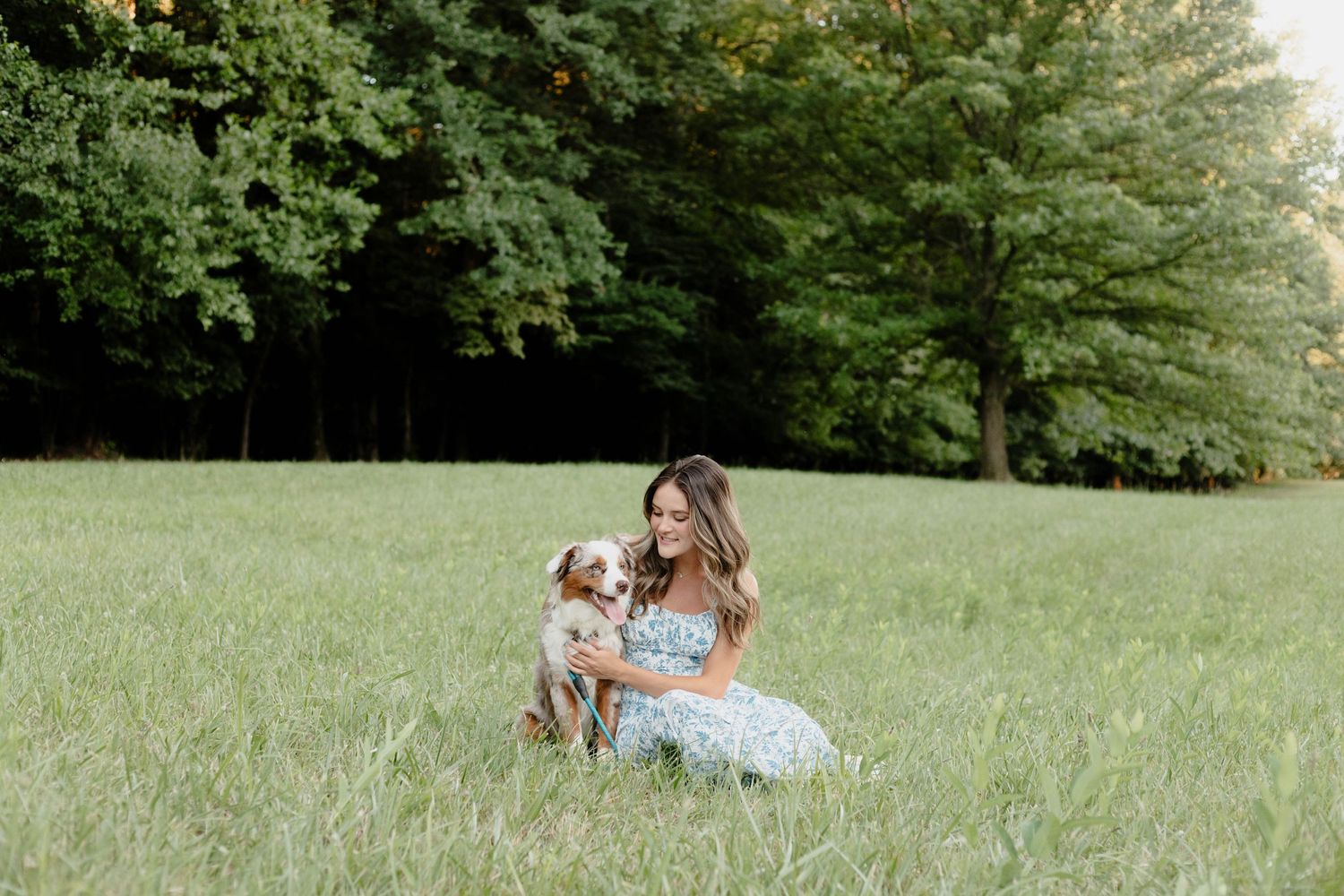 A sweet moment between a person in a floral dress and their Australian Shepherd dog in a sunny green field.