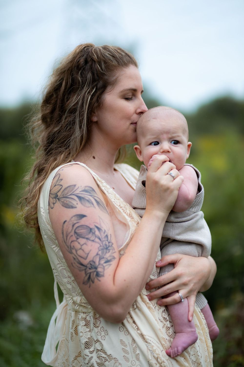 A person with floral tattoos holds and kisses a baby while wearing a cream-colored dress outdoors.