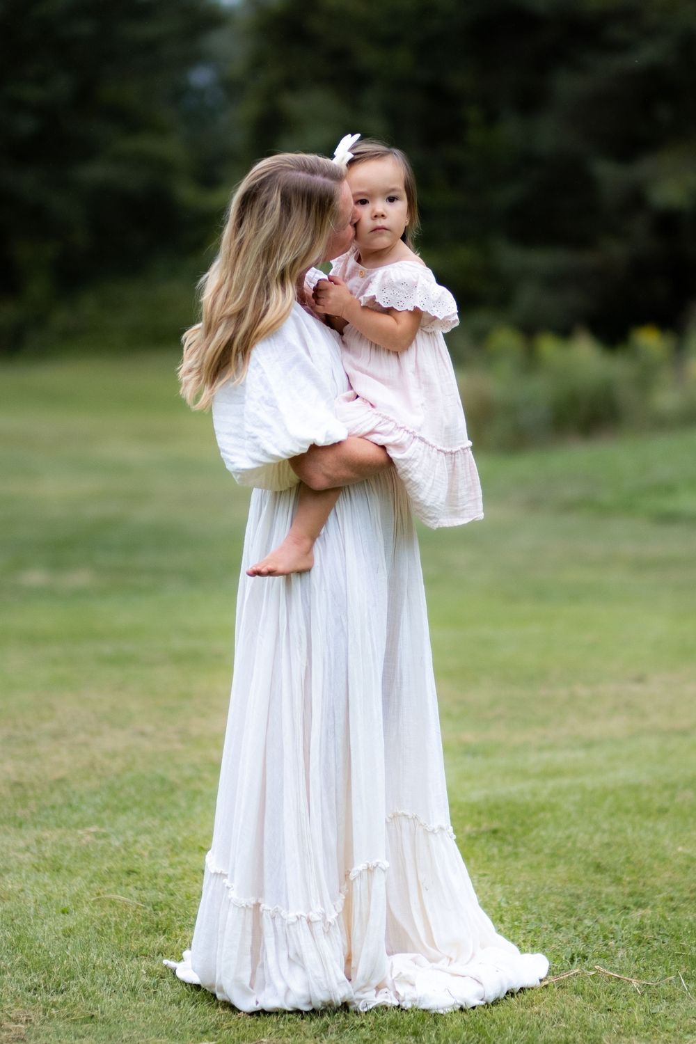 A person in a flowing white dress holds a small child in a garden setting.