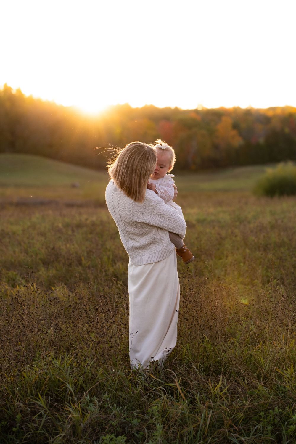 A person in white holds a baby against a golden sunset in a rural field.