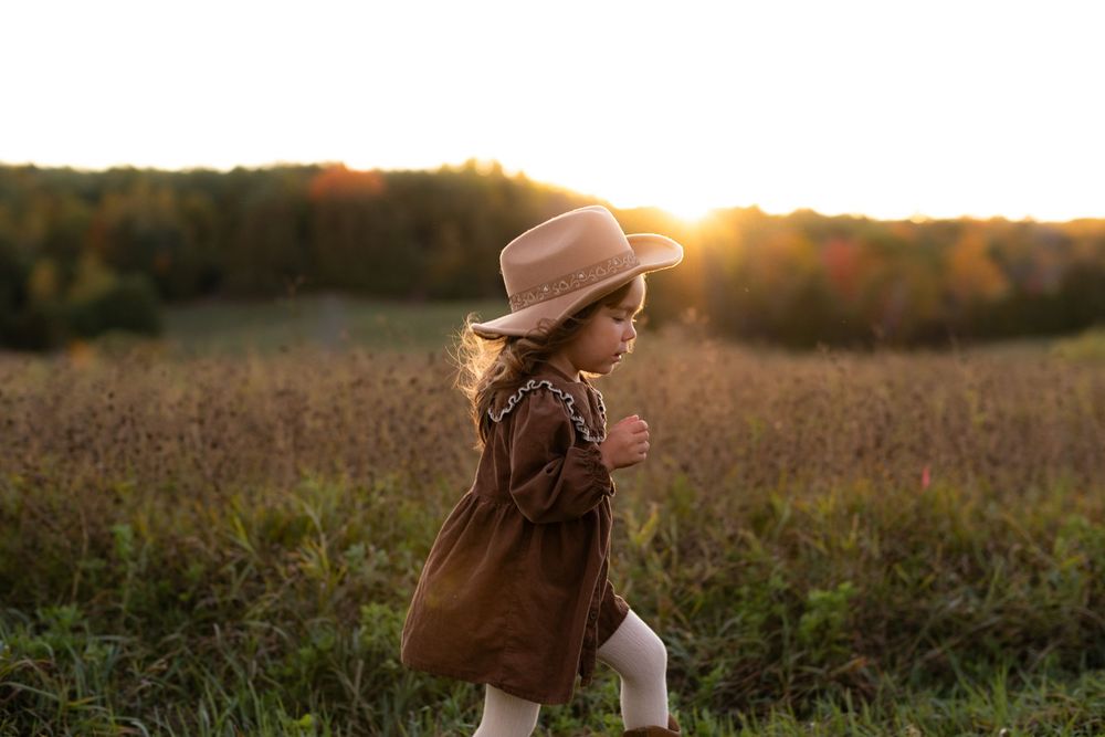 A child in a brown coat and hat stands in a sunlit field during golden hour.