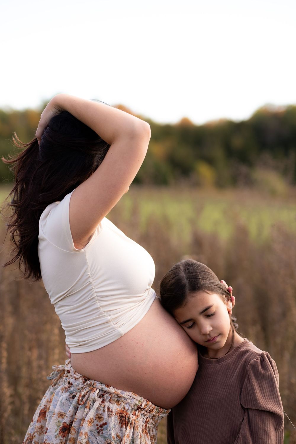 A tender moment between a pregnant woman in a white top and floral skirt with a child in a brown sweater outdoors.