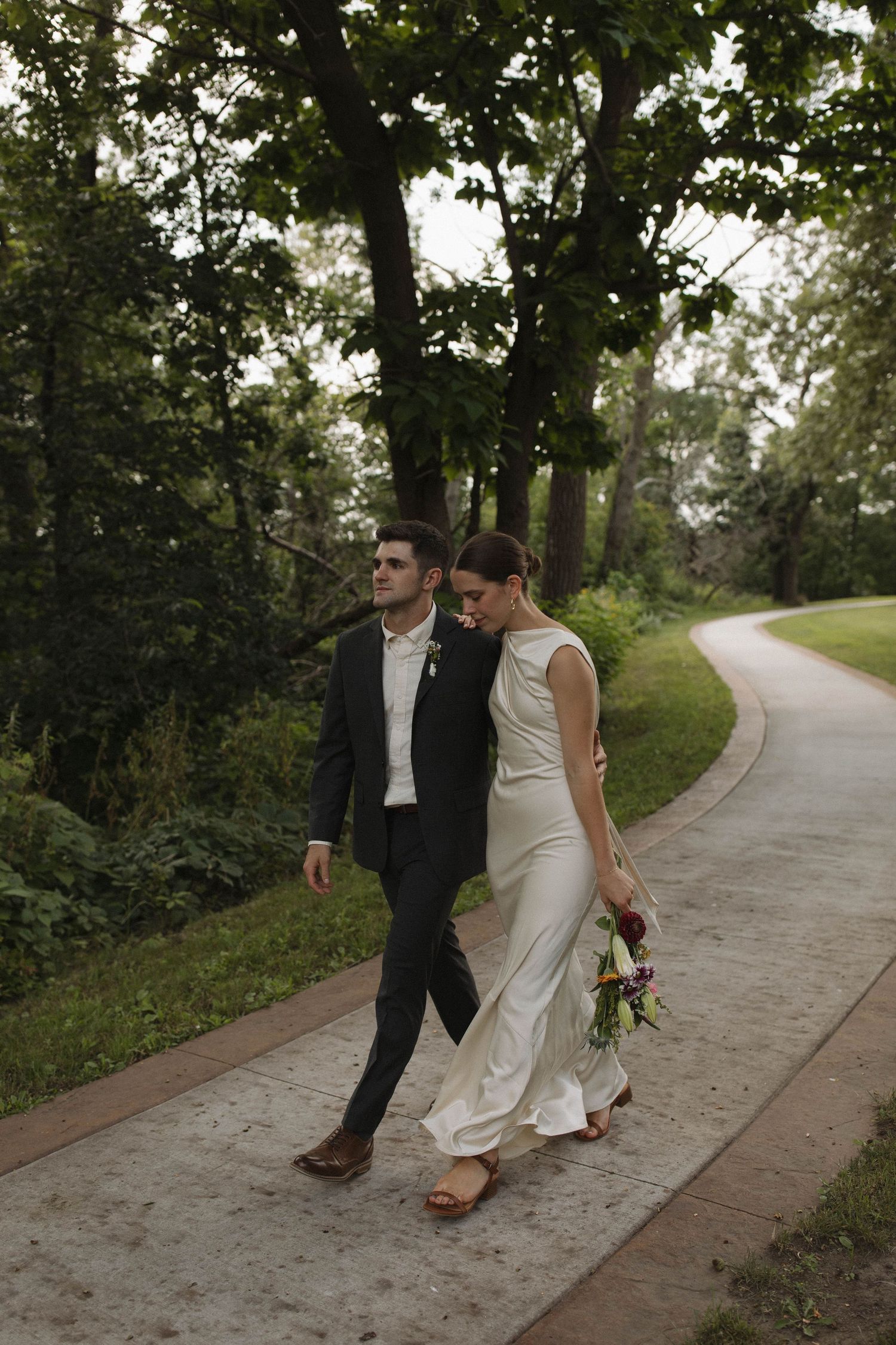 A couple walking together on a tree-lined path, the bride in a white dress and groom in a dark suit.