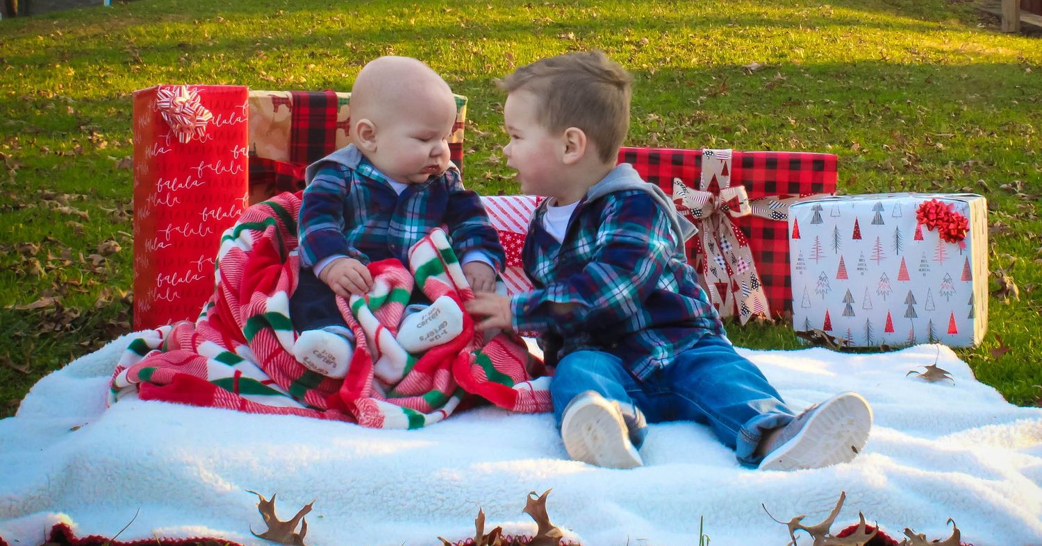 Two young children sitting on a blanket surrounded by wrapped Christmas presents.