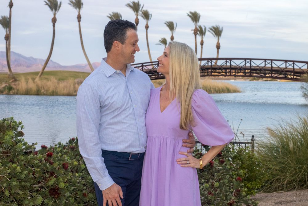 A couple in coordinated blue and lavender outfits smiles at each other near a scenic waterway with palm trees.