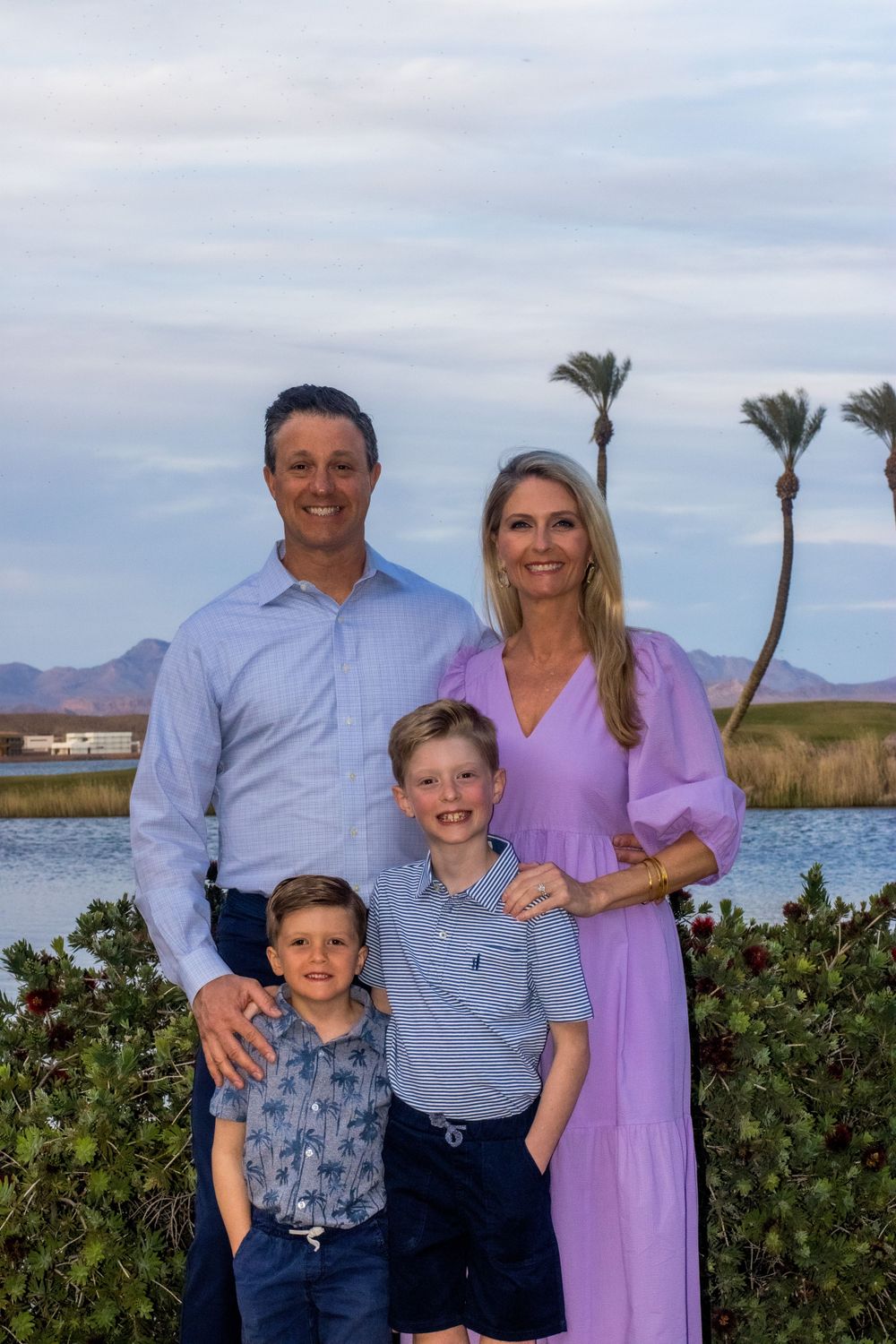 Family of four smiling together outdoors near a lake with palm trees at sunset.