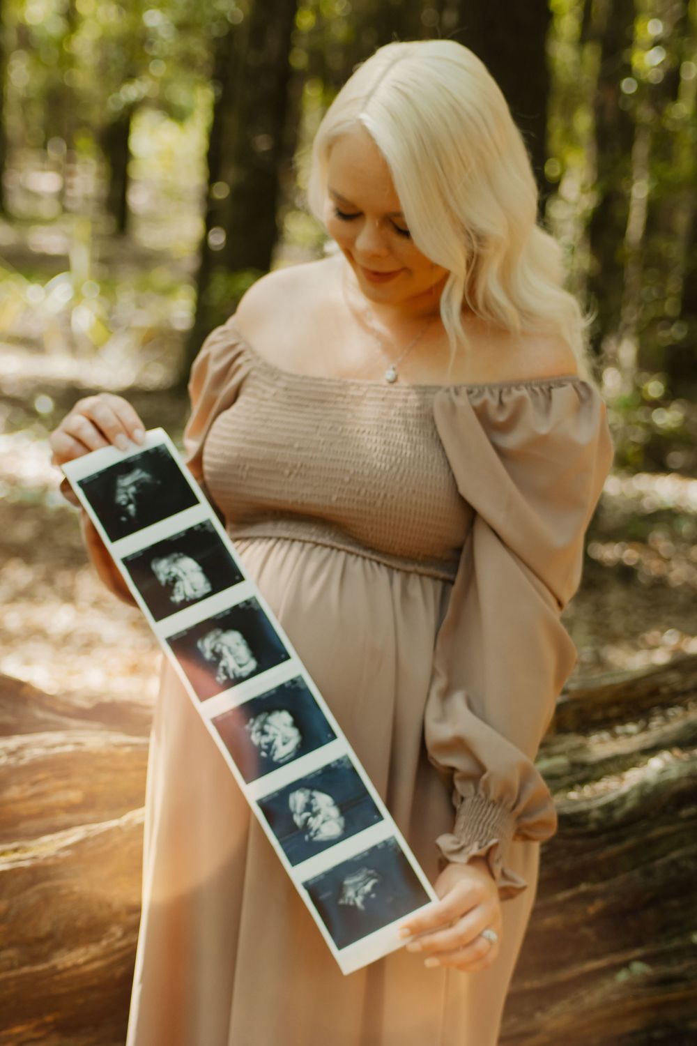 Expectant mother in a beige off-shoulder dress holds ultrasound photos while standing in a sunlit forest setting.