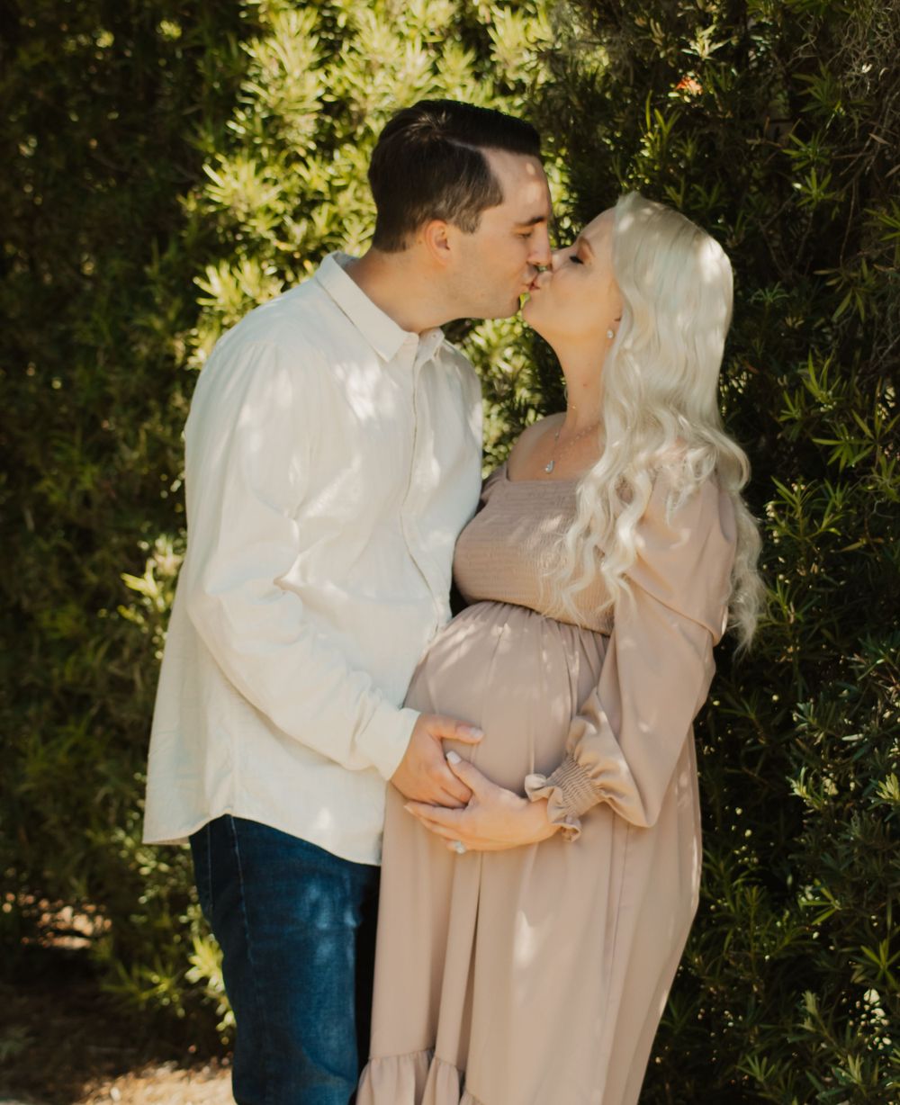 A couple shares an intimate embrace in a garden setting with sunlit foliage in the background.