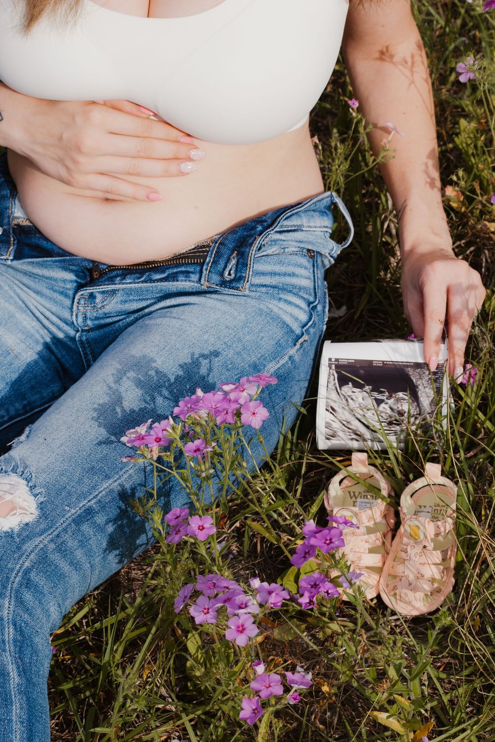 Pregnant woman in jeans holds ultrasound photos beside baby sandals and purple wildflowers in grass.