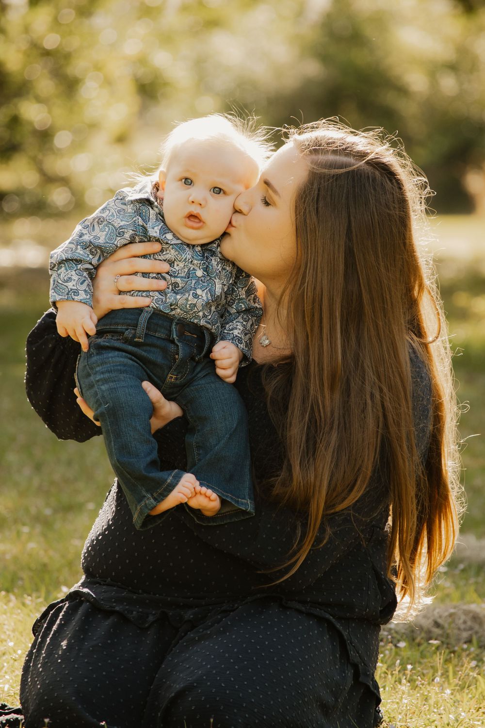 Loving mother with long brown hair kissing her wide-eyed baby dressed in a floral shirt in a sunny park.