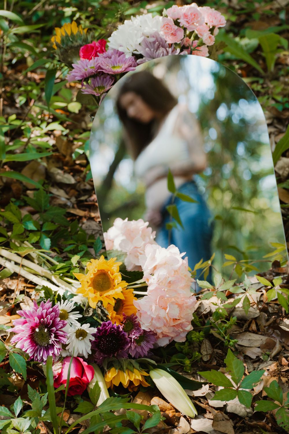 Arched mirror surrounded by colorful flowers on forest floor reflects pregnant woman holding belly outdoors.