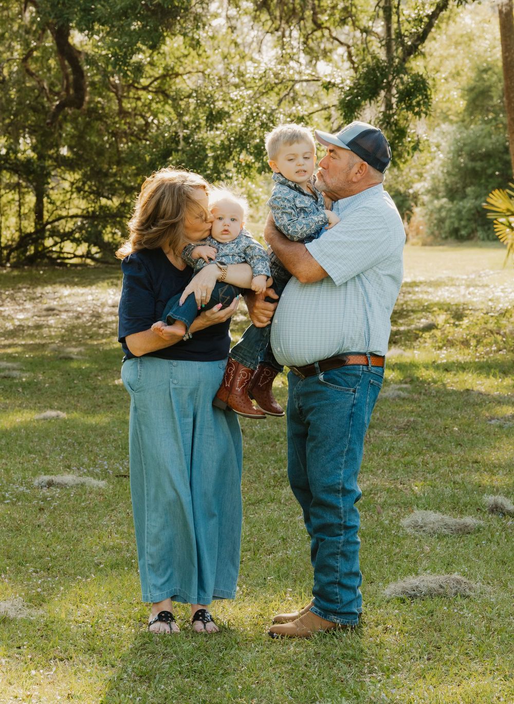 Grandparents kissing two young grandchildren outdoors in a green park, all dressed in casual coordinated outfits.