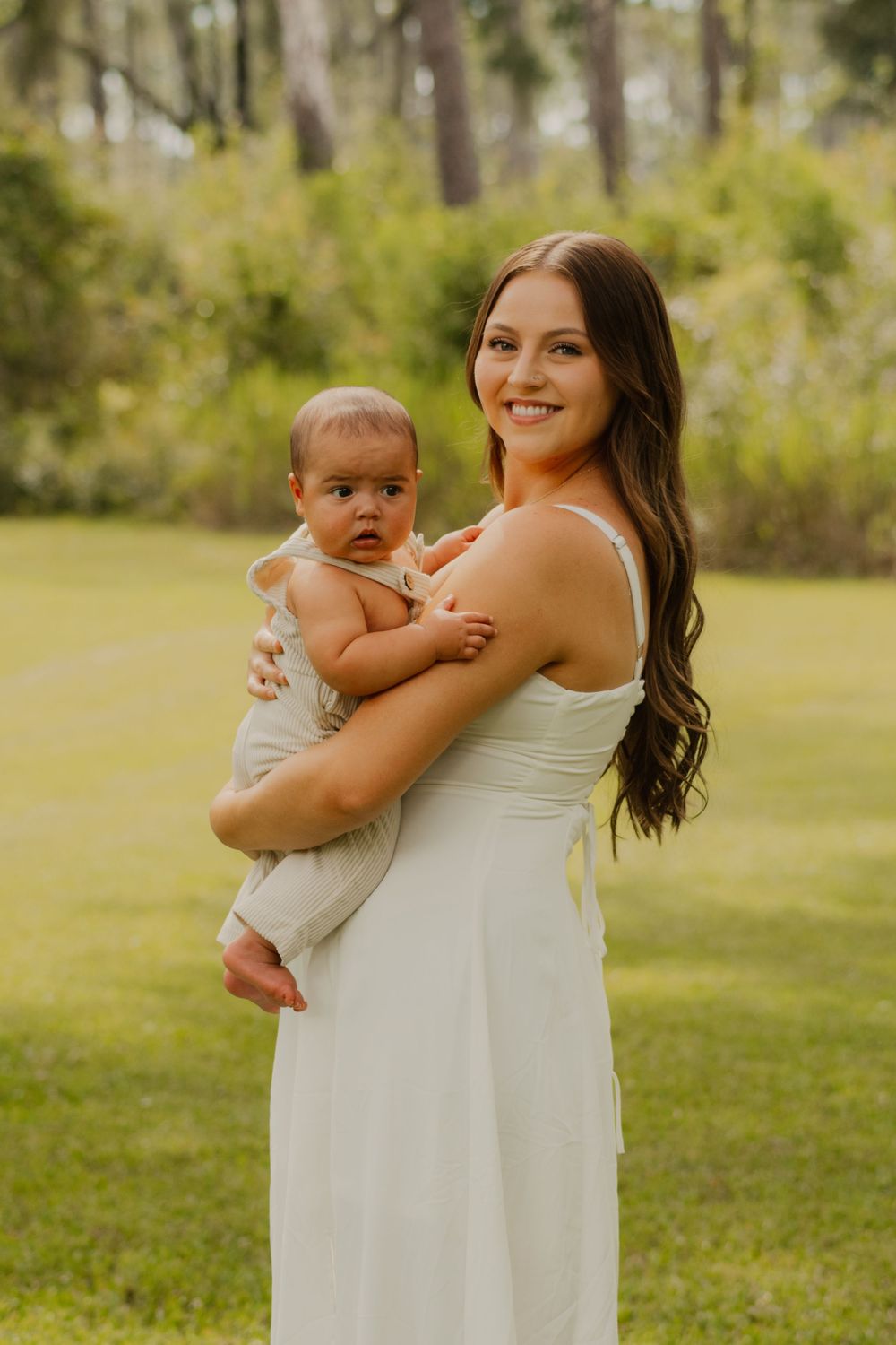A mother in a white dress holds a baby outdoors in a sunny park setting with trees in the background.