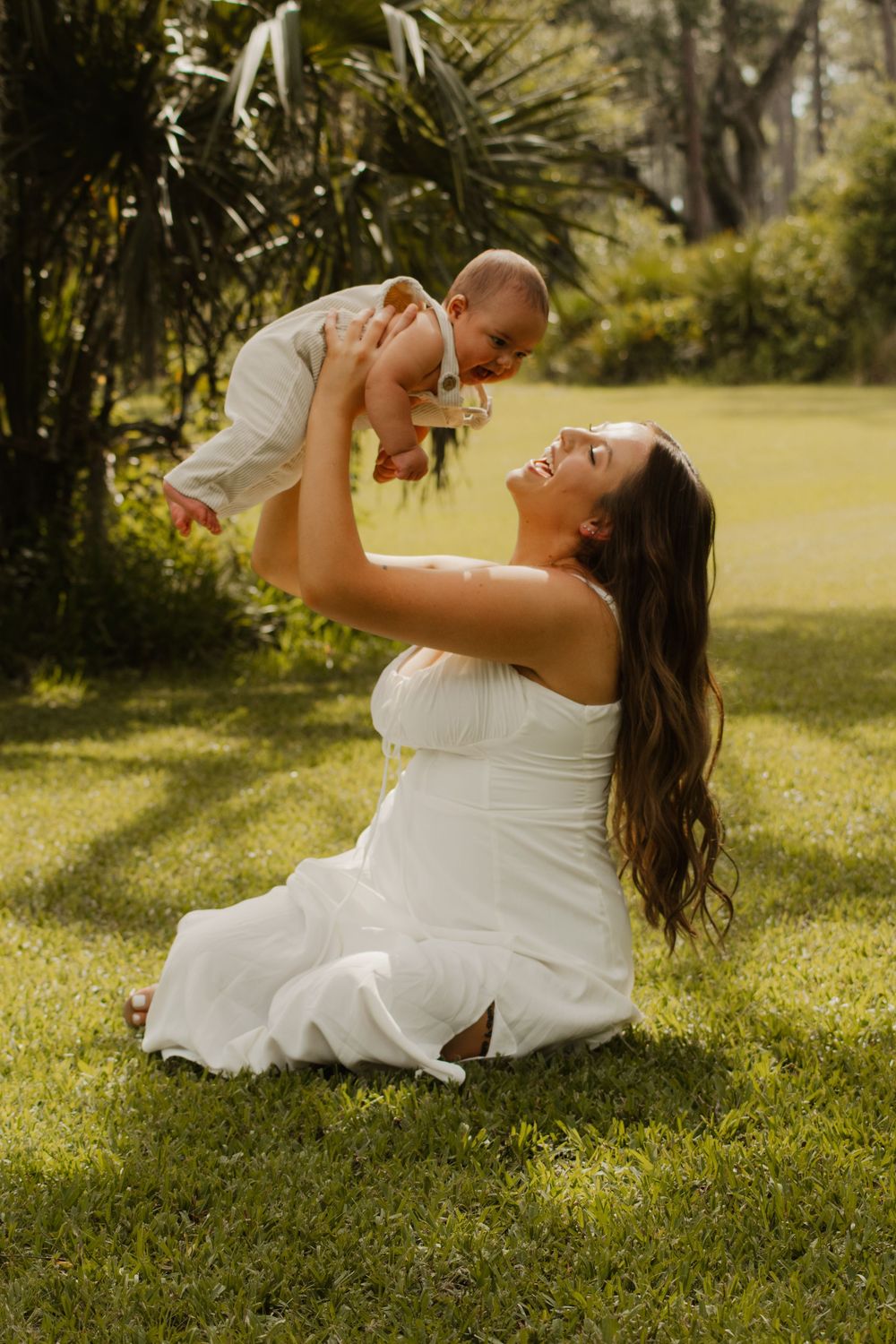 A mother and son in white clothing shares a joyful moment together on a sunlit grassy lawn during a romantic photoshoot.