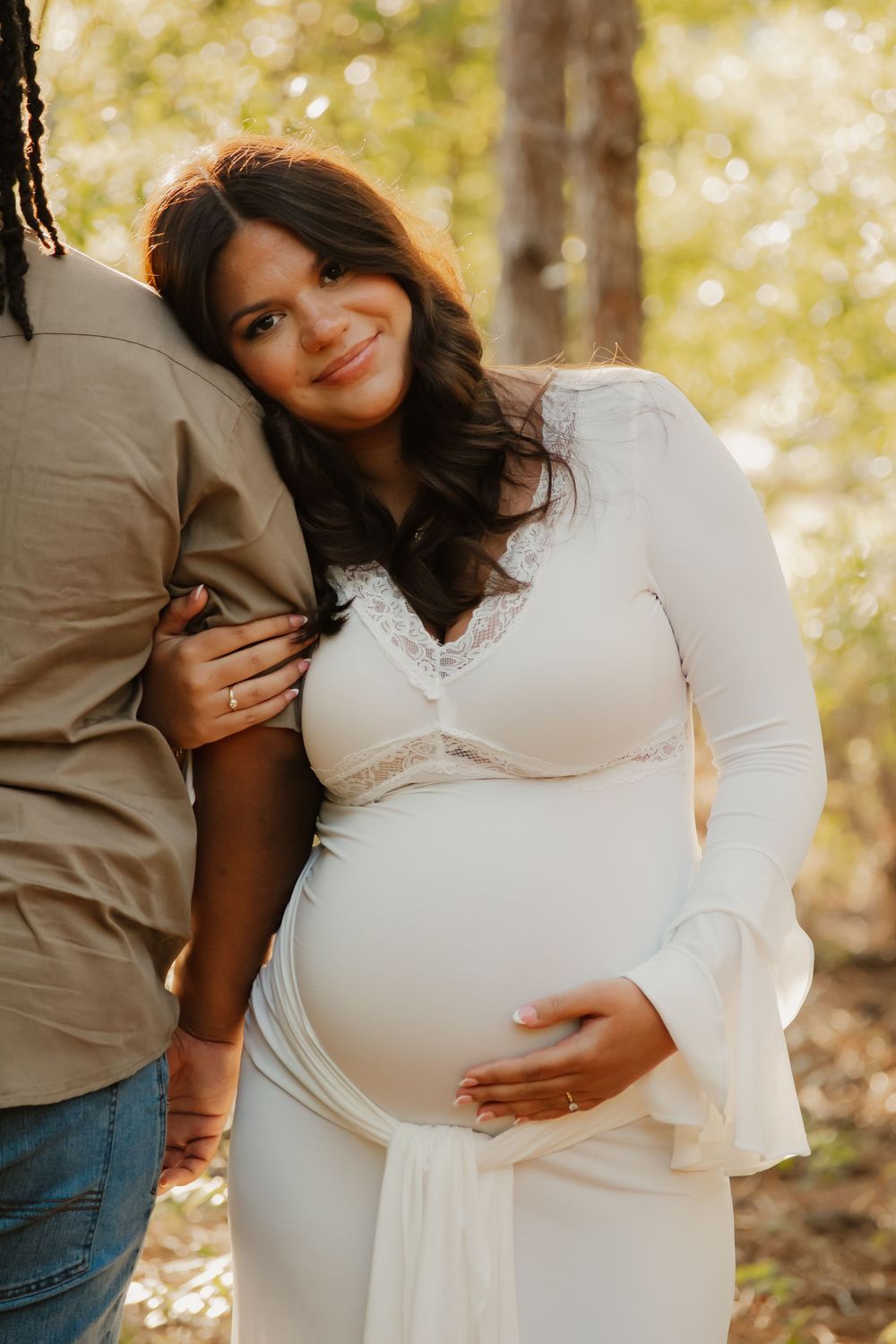 Pregnant woman in white lace dress leans against partner outdoors in sunlit forest during maternity photo session.