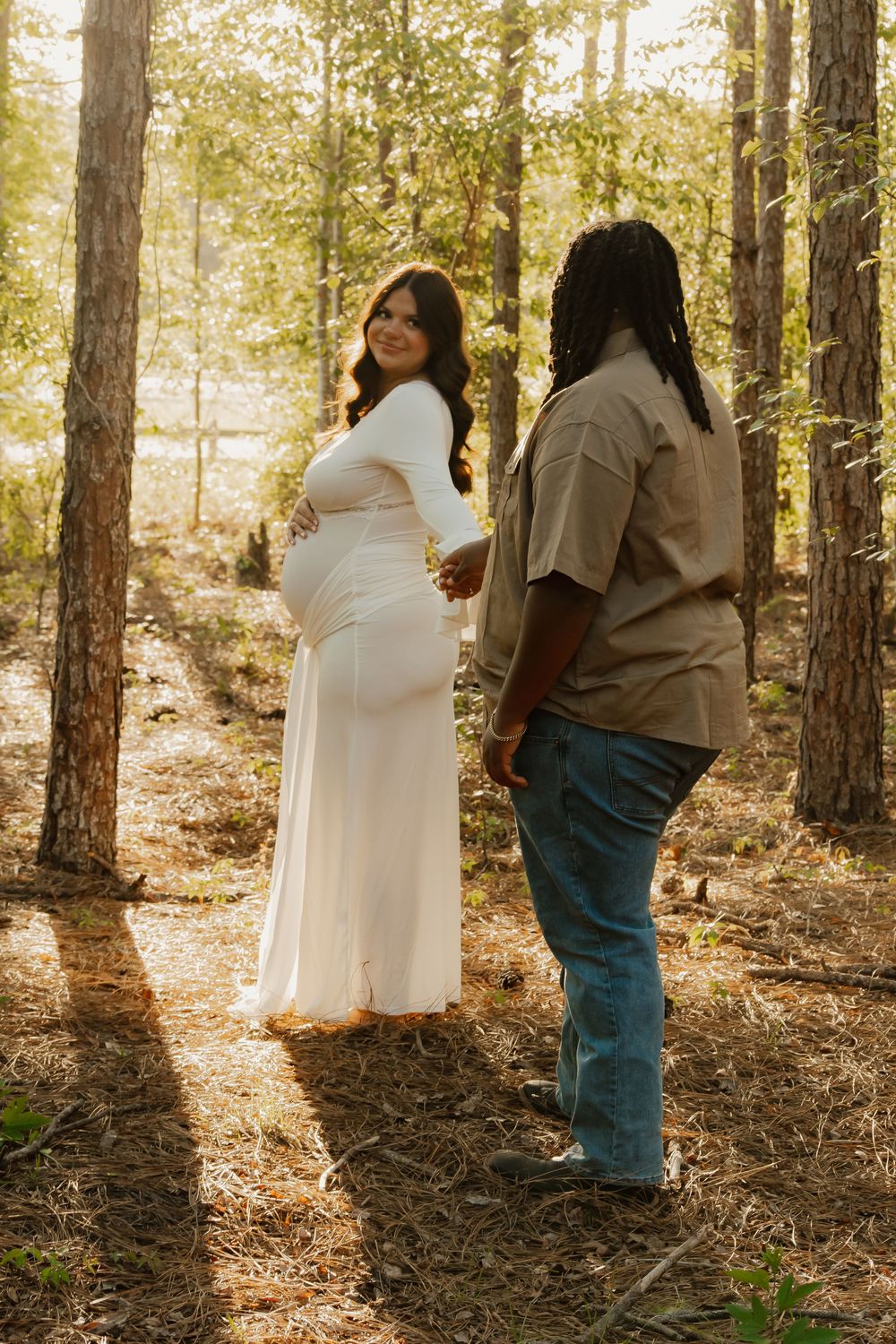 Interracial couple holds hands in sunlit pine forest during outdoor maternity photoshoot, woman wearing white dress.