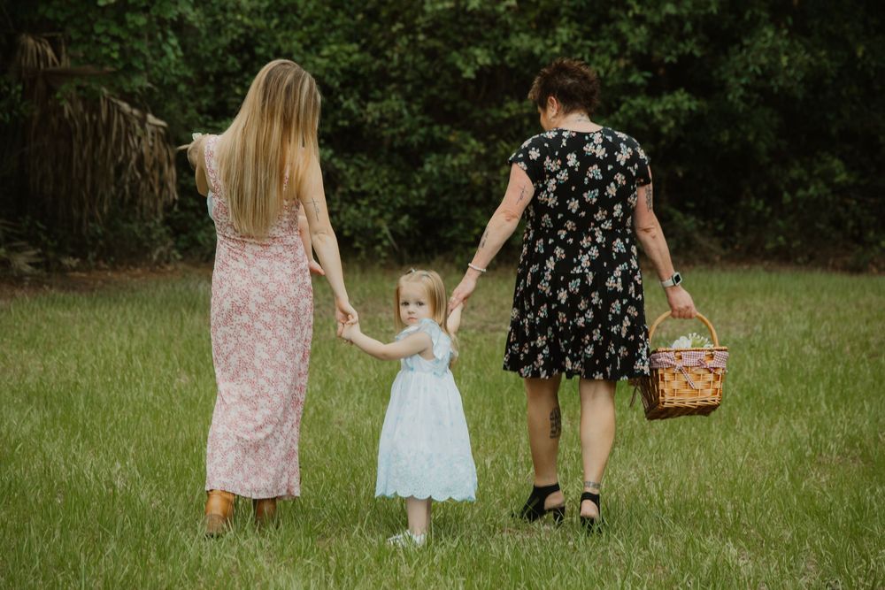 Two women and a young girl in light blue dress walk hand-in-hand across green grass carrying a picnic basket.