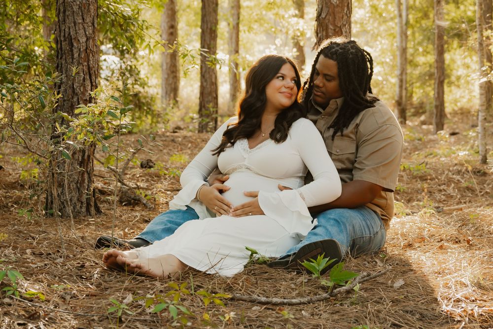 Couple sits together on forest floor during maternity session, man embraces pregnant woman in white dress.