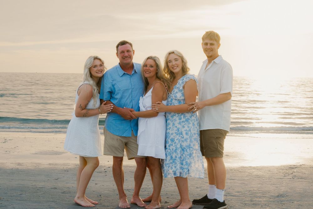 Family of five smiling together on a sandy beach at golden hour sunset with ocean waves in the background.