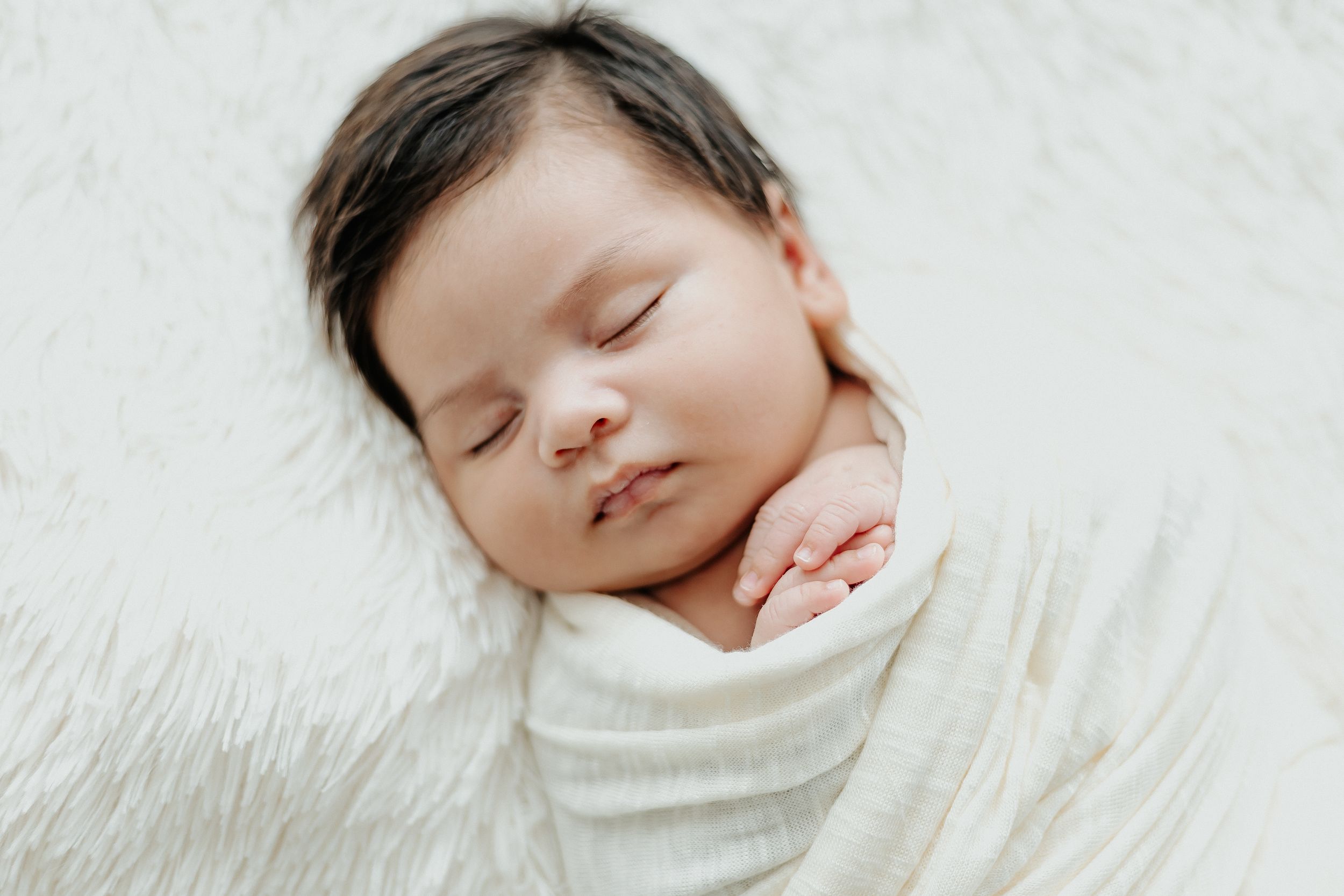 Newborn baby sleeping peacefully in white wrap against light background.