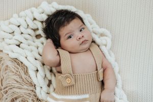 Baby wearing tan overalls lying on chunky knit blanket with peaceful expression.